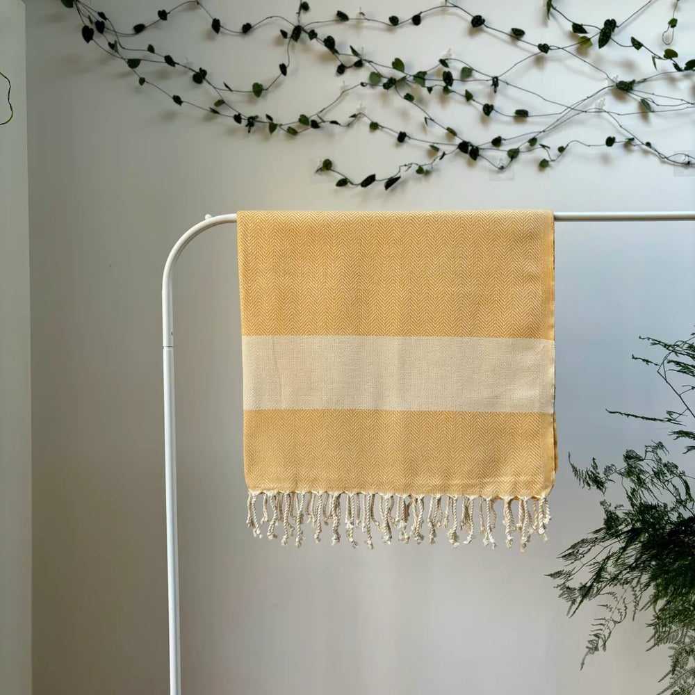 100% organic cotton herringbone patterned yellow and white Turkish Towel hanging on a clothes rack surrounded by plants on a white background