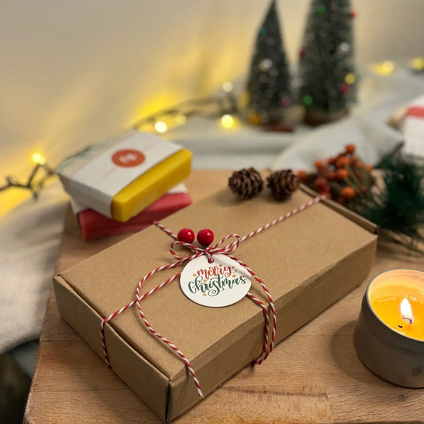 A christmas gift box with two organic soaps, a lit candle and christmas lights and decorations in the background