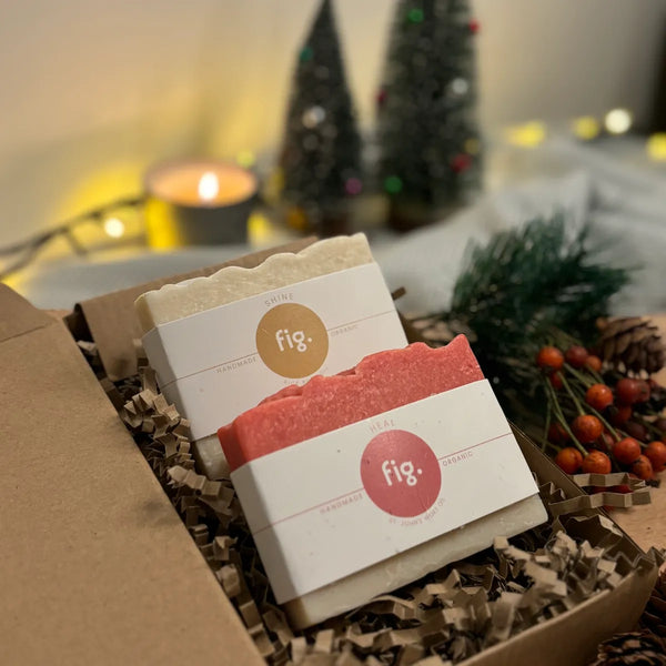 Two organic soaps on display in their gift box with a lit candle and christmas decorations in the background