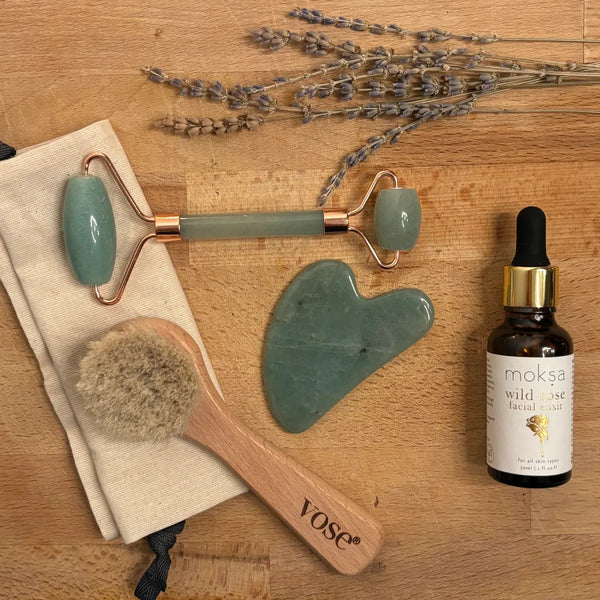 A horse hair face brush with a handle and cotton pouch, aventurine face roller and gua sha tool, and a facial elixir in a bottle on a table with dry lavender detail.