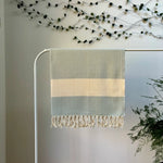 100% organic cotton herringbone patterned light blue and white Turkish Towel hanging on a clothes rack surrounded by plants on a white background