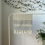 100% organic cotton grey Turkish Towel with white stripes hanging on a clothes rack surrounded by plants on a white background