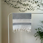 100% organic cotton white Turkish Towel with navy blue stripes hanging on a clothes rack surrounded by plants on a white background