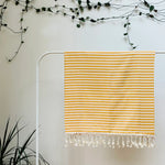 100% organic cotton stripy yellow and white Turkish Towel hanging on a clothes rack surrounded by plants on a white background