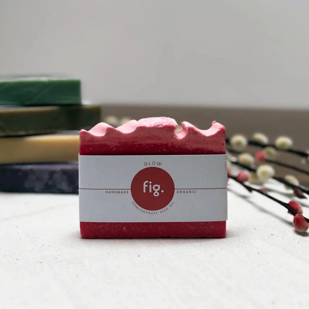 A pink handmade soap bar with a white paper label reading 'fig.' and 'GLOW', placed on a light surface with blurred stacked bars and decorative branches in the background.