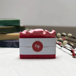 A pink handmade soap bar with a white paper label reading 'fig.' and 'GLOW', placed on a light surface with blurred stacked bars and decorative branches in the background.
