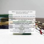 A green and white dual-purpose hair and skin cleansing bar is shown on a light surface with a text overlay detailing its natural ingredients and benefits. In the background, stacked green and gold cosmetic tubes are visible along with decorative beads.