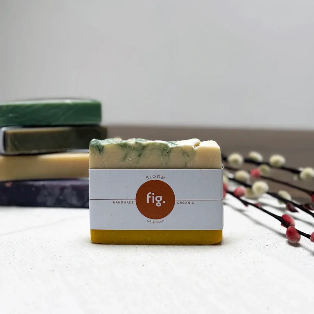 A single handmade bar of soap from the brand fig. stands in the center on a light surface, with other stacked soap bars blurred in the background and decorative branches on the right.