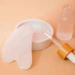 Pink gua sha tool, white bowl, and clear dropper bottle on a peach background