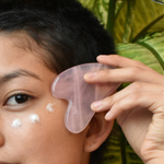 Person using a pink gua sha tool on their face with a natural background