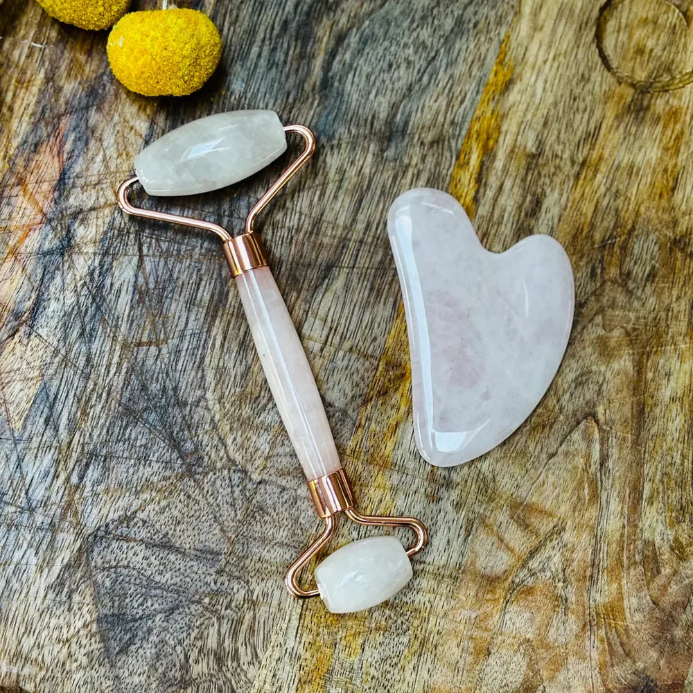 Rose quartz roller and gua sha tool on a wooden surface with yellow flowers.