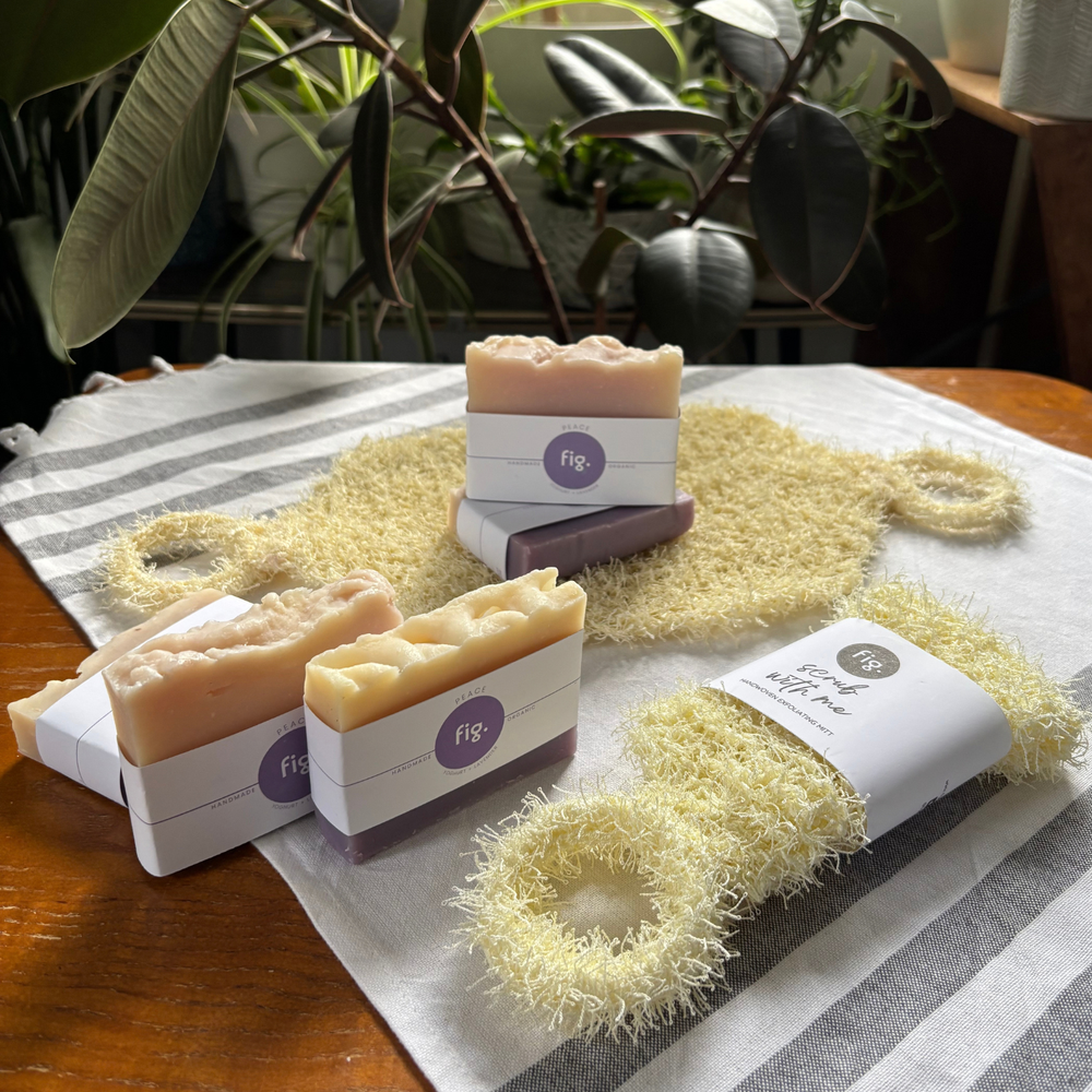 Three handmade soap bars and three natural loofah scrubber rings arranged on a striped cloth on a wooden table, with potted plants in the background. The soaps are in white paper sleeves with a purple circular logo reading 'fig', and one sleeve also reads 'Coconut & Coconut Oil Soap'.