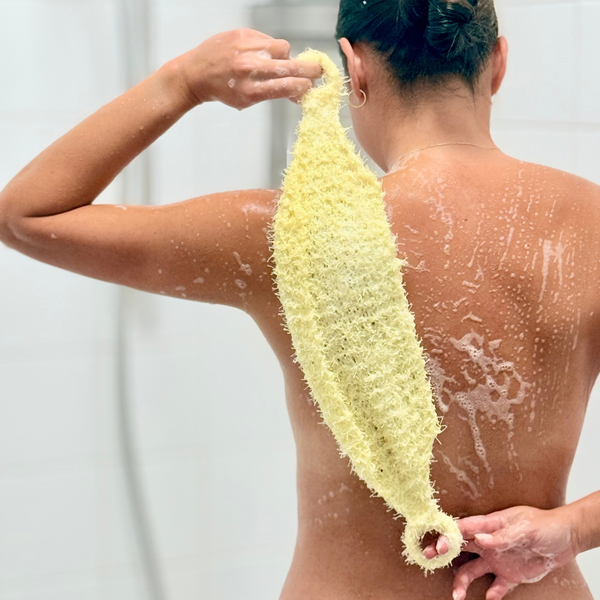 A person shown from behind in a shower, holding a pale yellow loofah sponge against their back. The person is wet with soap foam, and the loofah is being demonstrated for back scrubbing.