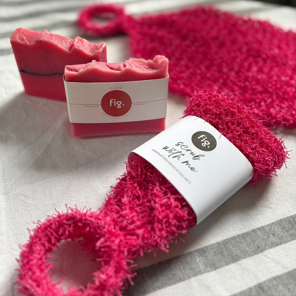 A close-up product shot showing a bright pink scrubber with a loop handle and a matching wrapped bar of soap on a light fabric surface. The soap wrapper shows the brand fig.