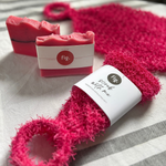 A close-up product shot showing a bright pink scrubber with a loop handle and a matching wrapped bar of soap on a light fabric surface. The soap wrapper shows the brand fig.
