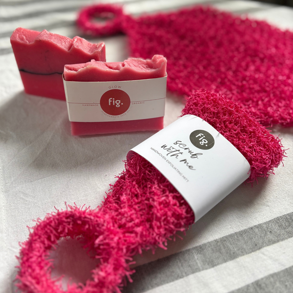 A close-up product shot showing a bright pink scrubber with a loop handle and a matching wrapped bar of soap on a light fabric surface. The soap wrapper shows the brand fig.