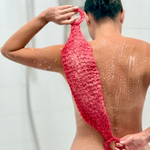 A woman from behind using a bright pink textured  shower mitt on her back in a shower setting.