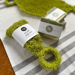 A lime green scrub ring with a white paper band labeled 'fig. scrub with me' lies on a white and gray striped cloth next to a wrapped bar of soap.