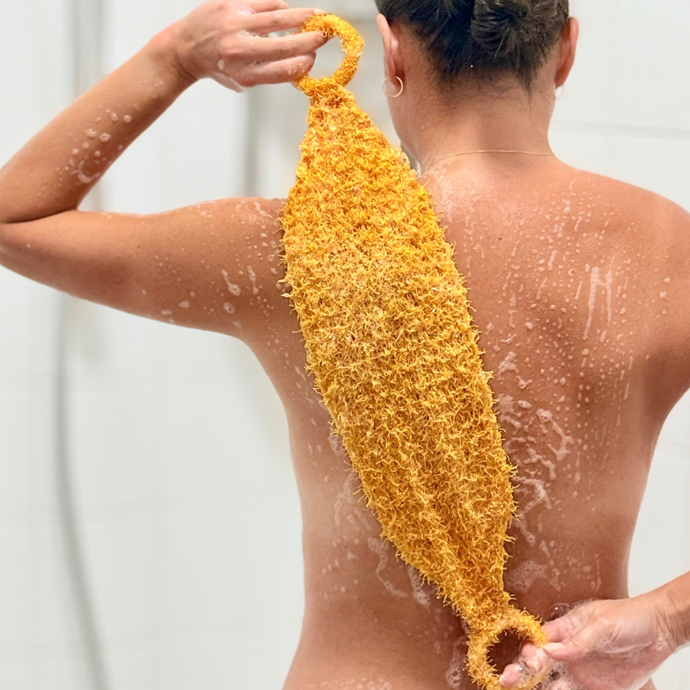 A person shown from behind in a shower, holding a yellow textured scrubber or loofah-style body scrubber against their back. Soap lather is visible on the skin, and the product is covered in yellow-orange scrubbing fibers.