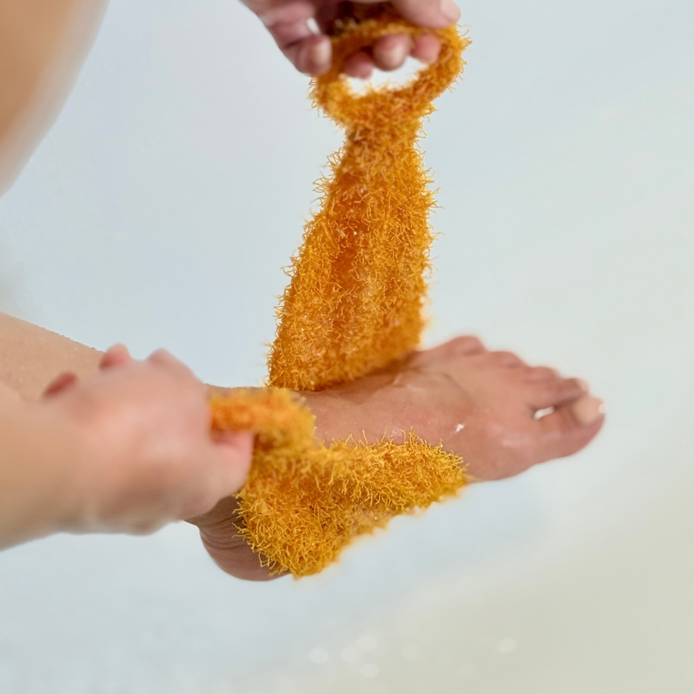 A close-up of a person using a bright orange, highly textured, looped foot scrubber on their foot, with water droplets visible on the skin against a clean, light background.