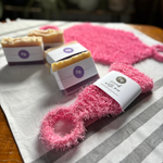 Pink fluffy scrubber mitts and packaged soap bars arranged on a striped cloth on a wooden table. The front scrubber mitt has a paper label reading Fig.