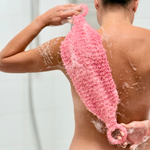 A person shown from the back in a shower, using a pink, porous scrubber or loofah-style body wash tool on their back. Soap lather is visible on the skin and the tool.