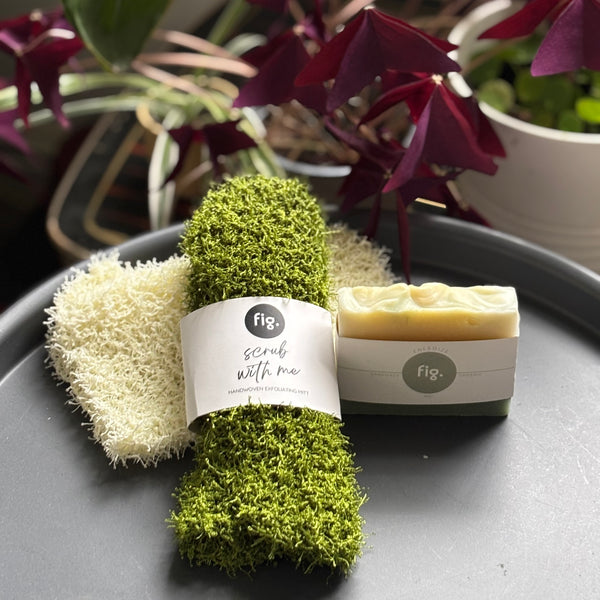Green shower scrub and a bar of soap on a gray surface with plants in the background