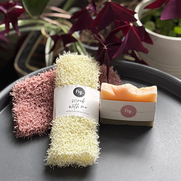 cream shower scrub with an organic radiance soap on a gray surface with plants in the background.