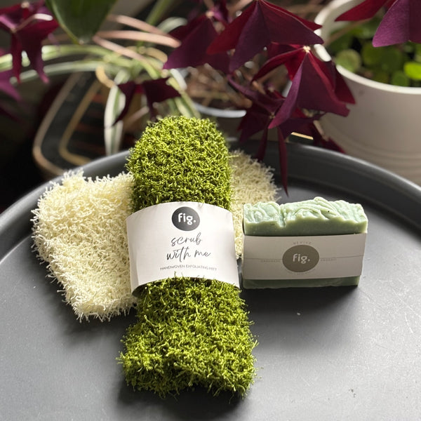 Green shower scrub and soap set on a gray surface with plants in the background.