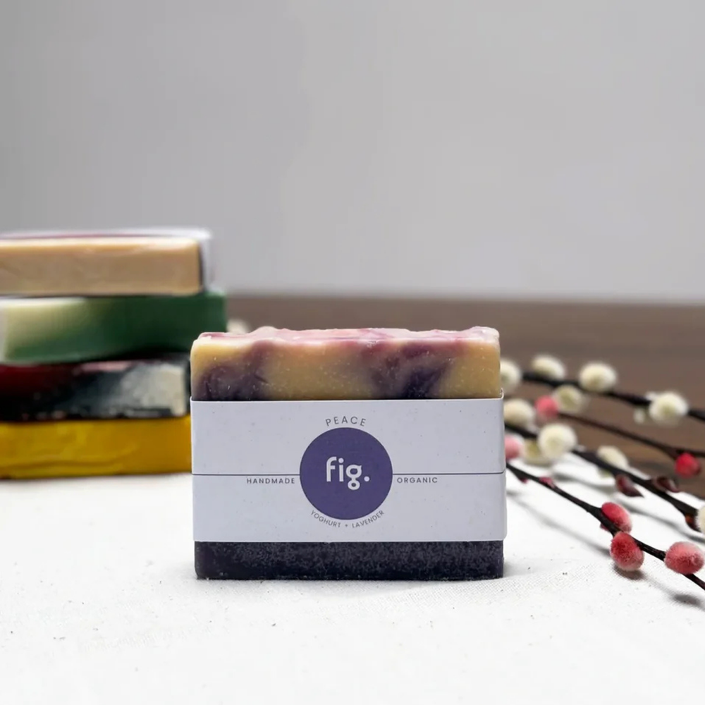 A single handmade soap bar wrapped in a white paper label stands upright on a light surface, with a stack of other colorful soaps blurred in the background and decorative branches on the right.