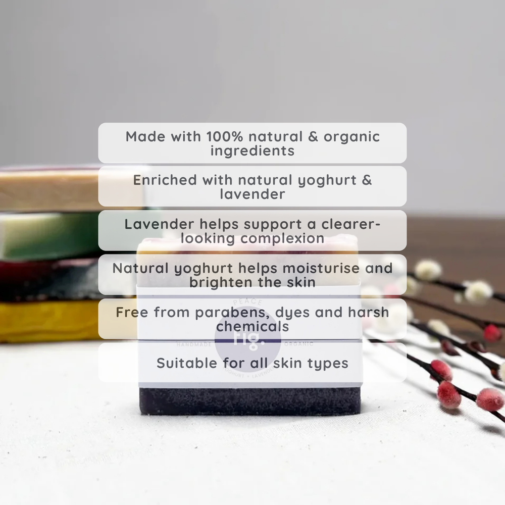 A rectangular bar soap product standing upright on a light surface, with a stack of similar bars blurred in the background. White text boxes overlay the center, describing natural ingredients, yogurt and lavender enrichment, skin benefits, and suitability for all skin types.