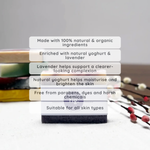 A rectangular bar soap product standing upright on a light surface, with a stack of similar bars blurred in the background. White text boxes overlay the center, describing natural ingredients, yogurt and lavender enrichment, skin benefits, and suitability for all skin types.