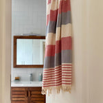 A striped bath towel with cream, dusty red, and slate blue horizontal bands hangs on a white door in a bathroom setting, with a sink and mirror visible in the soft-focus background.