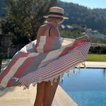 A woman standing by a swimming pool outdoors, holding open a large striped Turkish towel with tassels. The towel has alternating wide coral-red and off-white stripes with some red-and-white narrow stripes on one end.