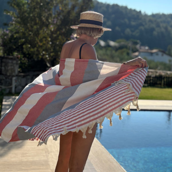 A woman standing by a swimming pool outdoors, holding open a large striped Turkish towel with tassels. The towel has alternating wide coral-red and off-white stripes with some red-and-white narrow stripes on one end.