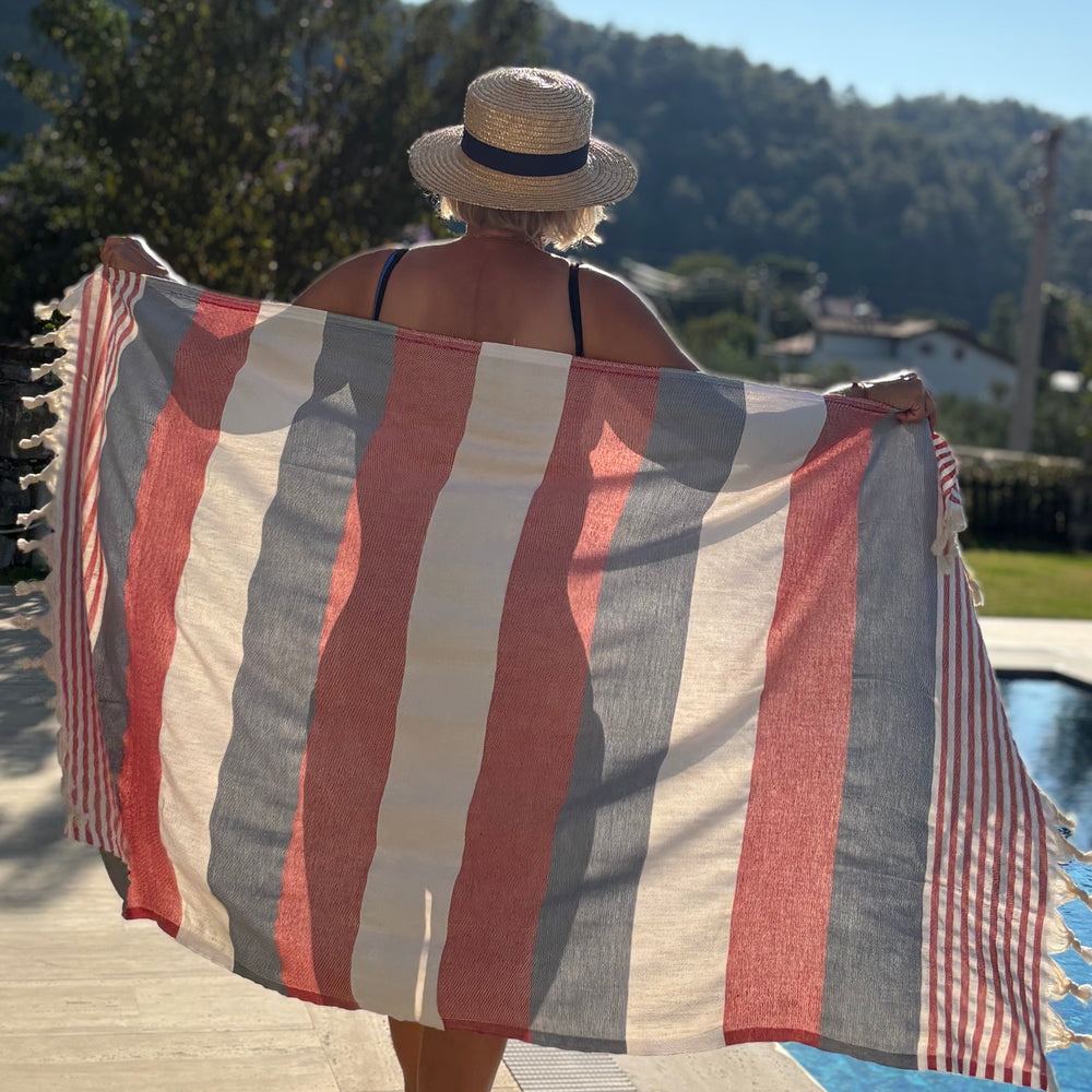 A woman stands by a pool holding up a large striped beach towel or sarong with red, cream, and gray vertical stripes and tasseled edges, with a sunny outdoor background.