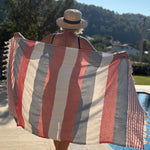 A woman stands by a pool holding up a large striped beach towel or sarong with red, cream, and gray vertical stripes and tasseled edges, with a sunny outdoor background.