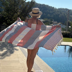 A woman stands by a swimming pool holding up a large striped beach towel. The towel has wide stripes in dusty pink, light gray, and white, with a section of red and white stripes and fringed edges.