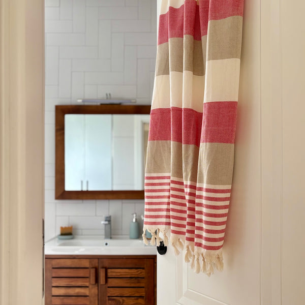 A striped Turkish cotton towel with cream, beige, and coral pink blocks and tassels, hanging on a white door in a bathroom setting.