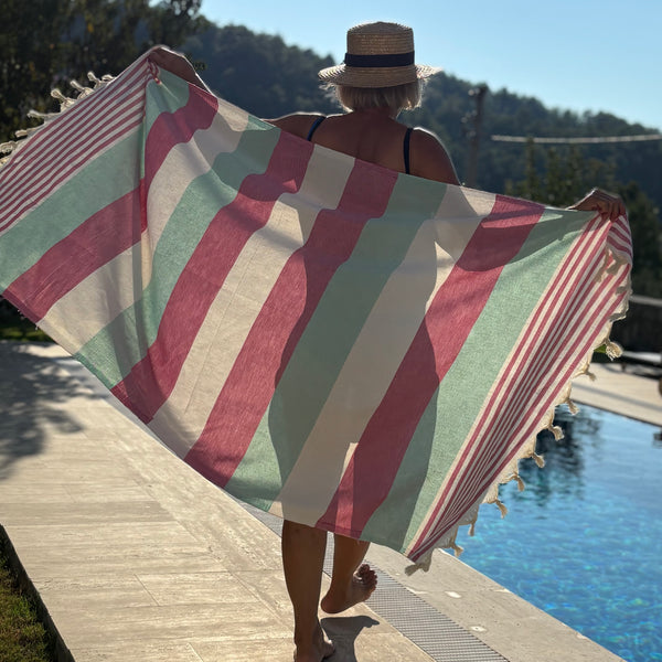 A woman standing by a swimming pool holds up a large striped beach towel or sarong with cream, dusty pink, and mint green vertical stripes and tassel trim, photographed outdoors with trees and hills in the background.