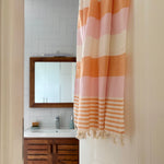 A striped bath towel with fringed tassels hanging in a doorway, showing a bathroom background with a sink, mirror, and wooden cabinet.