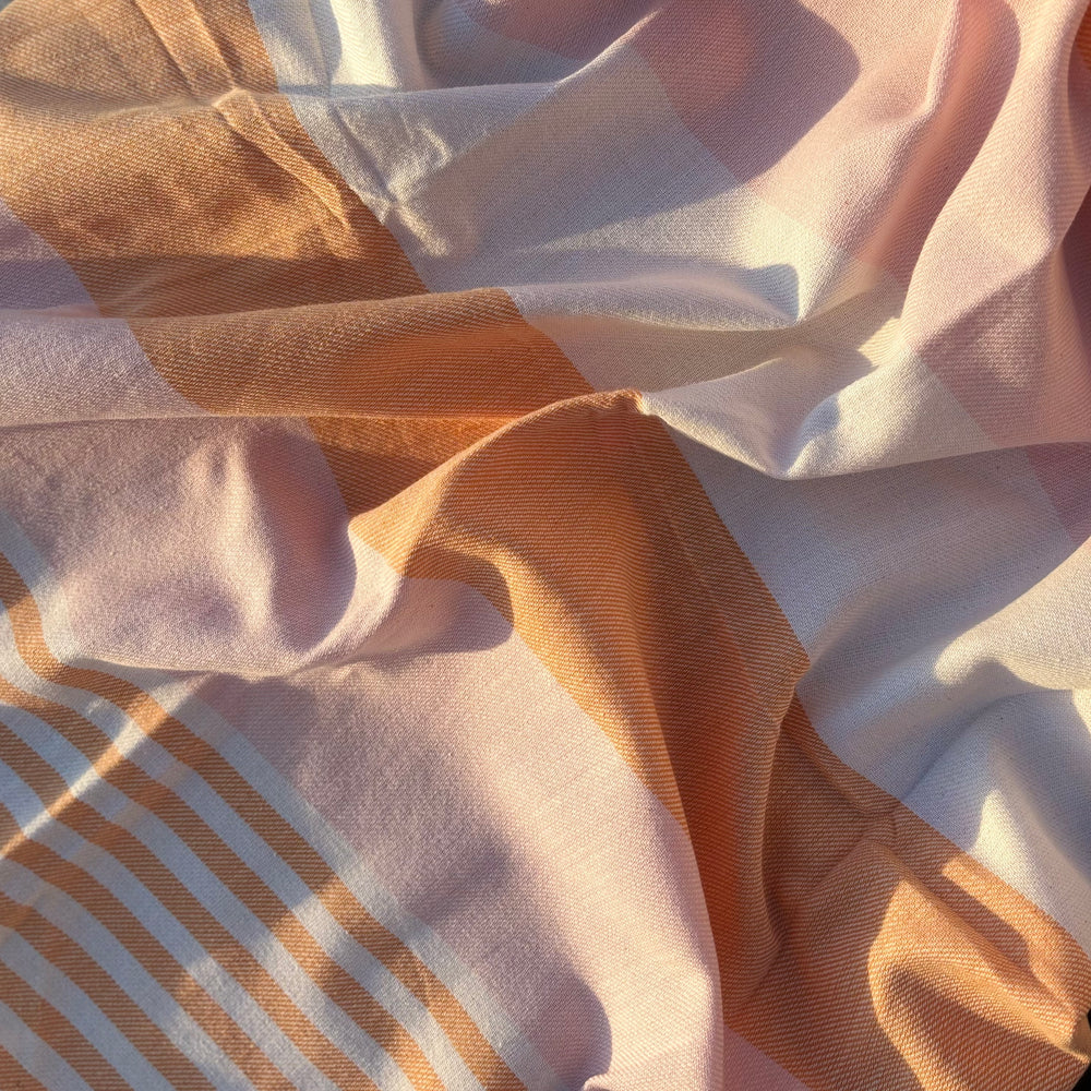 A close-up of draped fabric with a soft plaid pattern in warm peach, cream, and light pink tones, with sunlight casting shadows across the material.