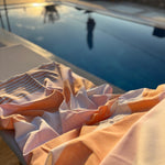 A close-up of a peach and white striped fabric accessory, likely a towel or blanket, draped on a poolside lounge chair with a swimming pool in the background.
