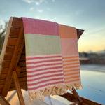 A striped cotton Turkish towel in pink, sage green, and peach-orange draped over a wooden outdoor lounge chair next to a swimming pool at sunset.