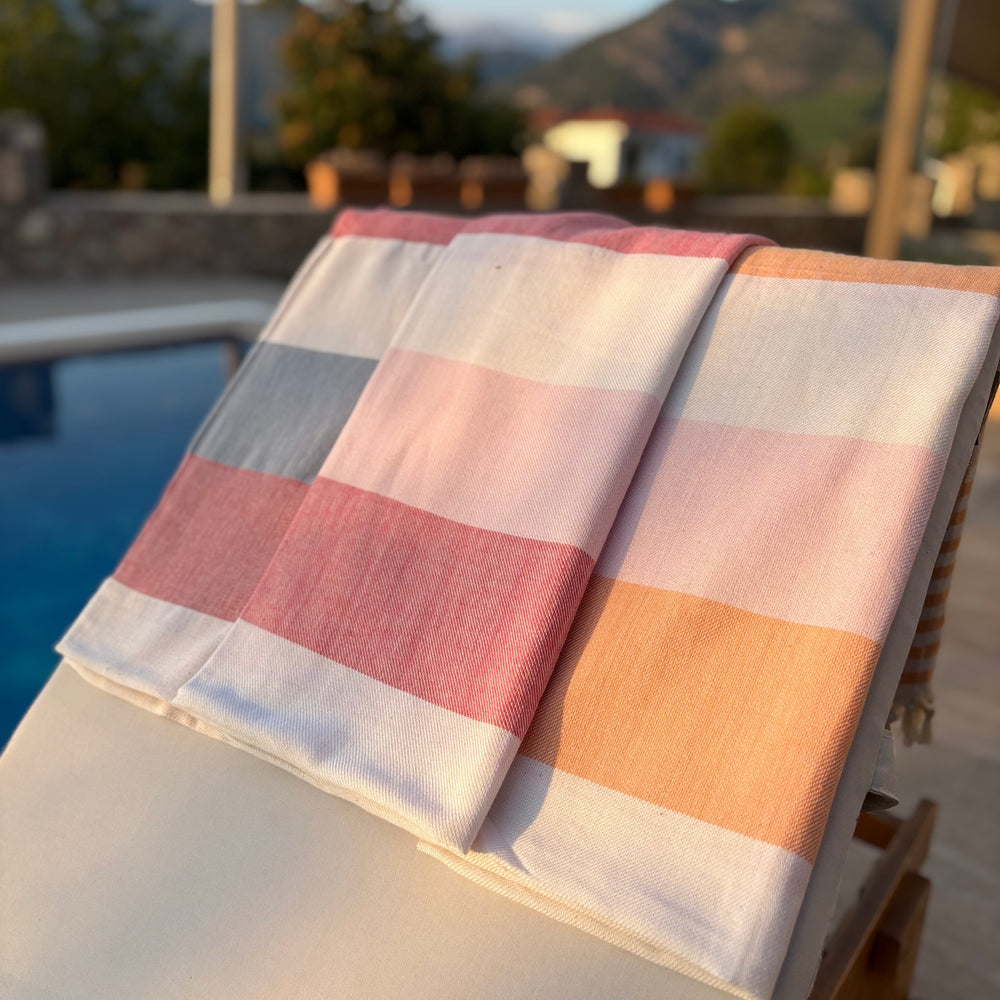 A folded striped beach towel with pink, peach, gray, and white stripes rests on a white lounge chair next to a swimming pool, with a blurred outdoor resort background.