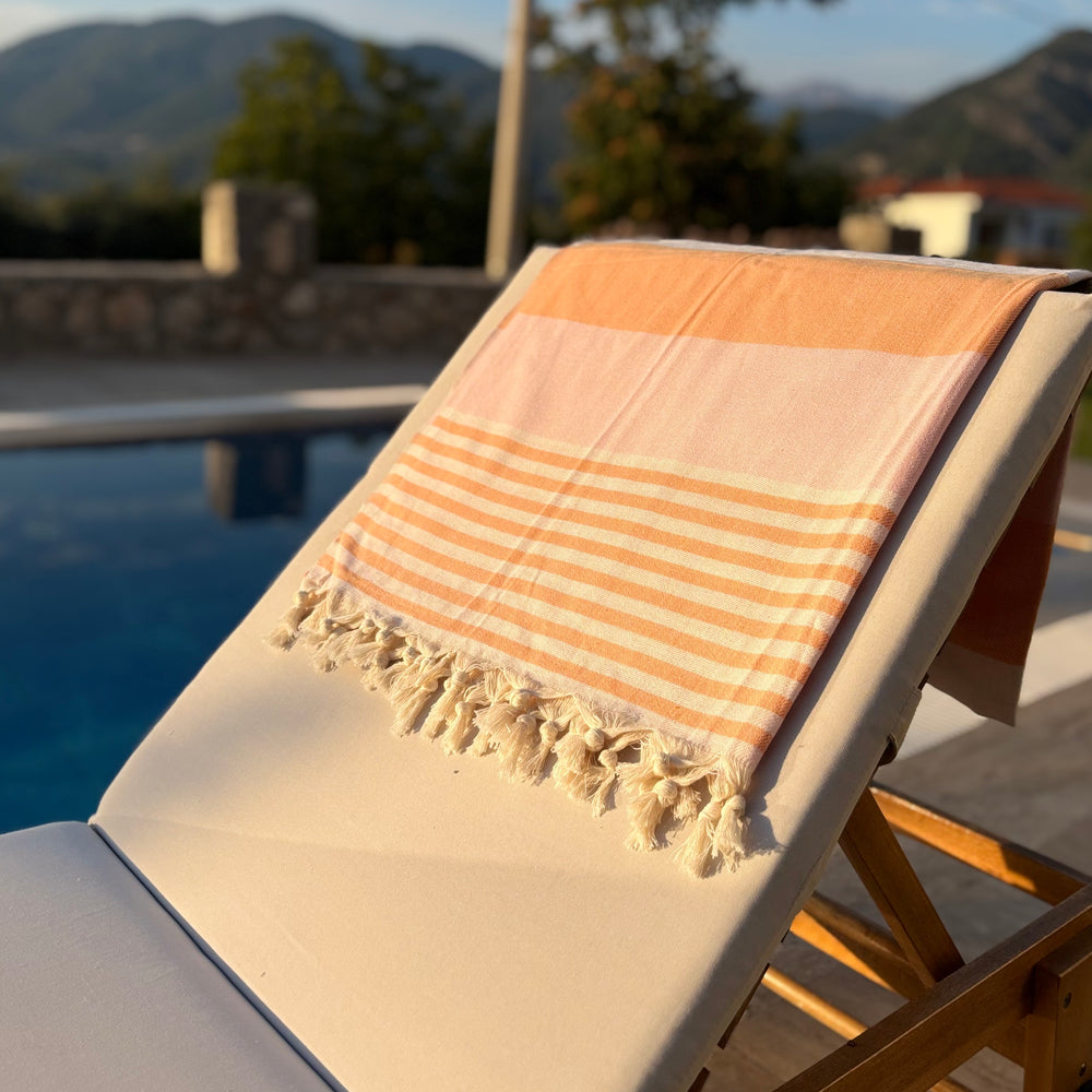A peach and cream striped Turkish cotton towel with fringed tassels draped over a white outdoor lounge chair next to a swimming pool, with a blurred mountain background.