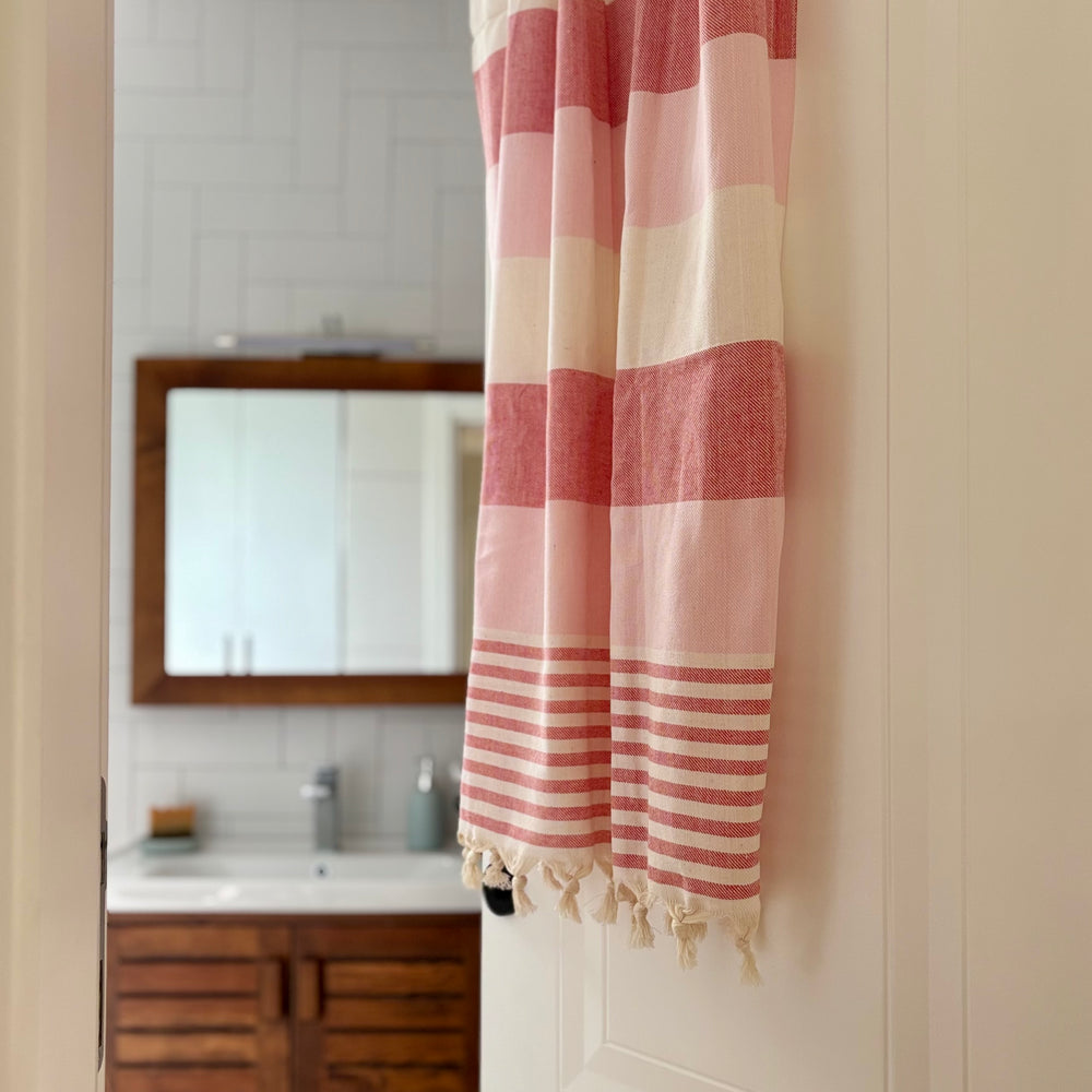 A pink and cream striped Turkish cotton towel with fringed tassels hanging on a white door in a bathroom setting.