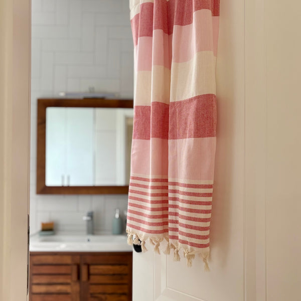 A pink and cream striped Turkish cotton towel with fringed tassels hanging on a white door in a bathroom setting.