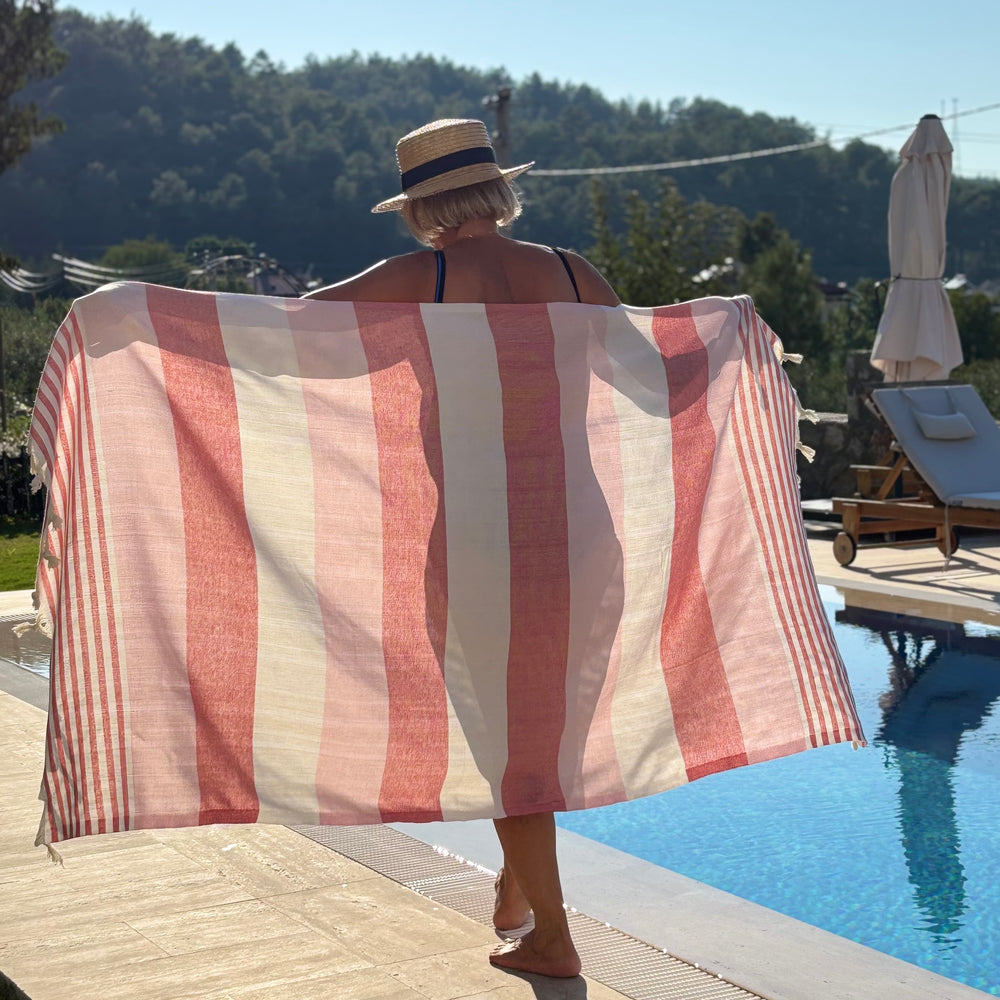 A woman standing by a swimming pool holds up a large striped beach towel or wrap with vertical bands of dusty pink, cream, and white. She is seen from behind, wearing a straw hat, with a sunny outdoor patio, lounge chair, and forested hill in the background.