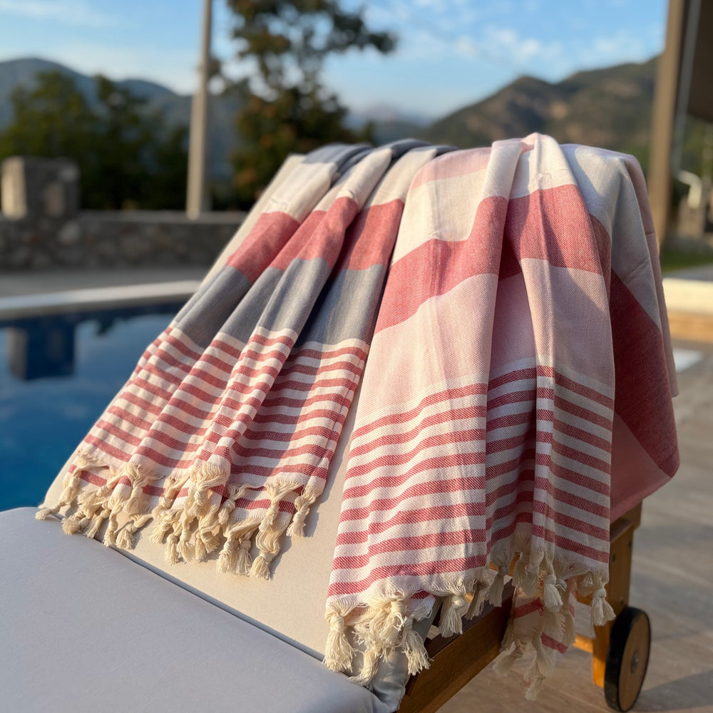 A striped Turkish cotton towel with cream tassels draped over a lounge chair outdoors near a pool and mountains.